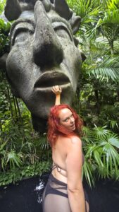 Nikki posing playfully beside a giant stone sculpture surrounded by tropical greenery in Tulum, Mexico