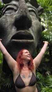 Nikki celebrating with her arms raised in front of a giant stone sculpture surrounded by tropical greenery in Tulum, Mexico